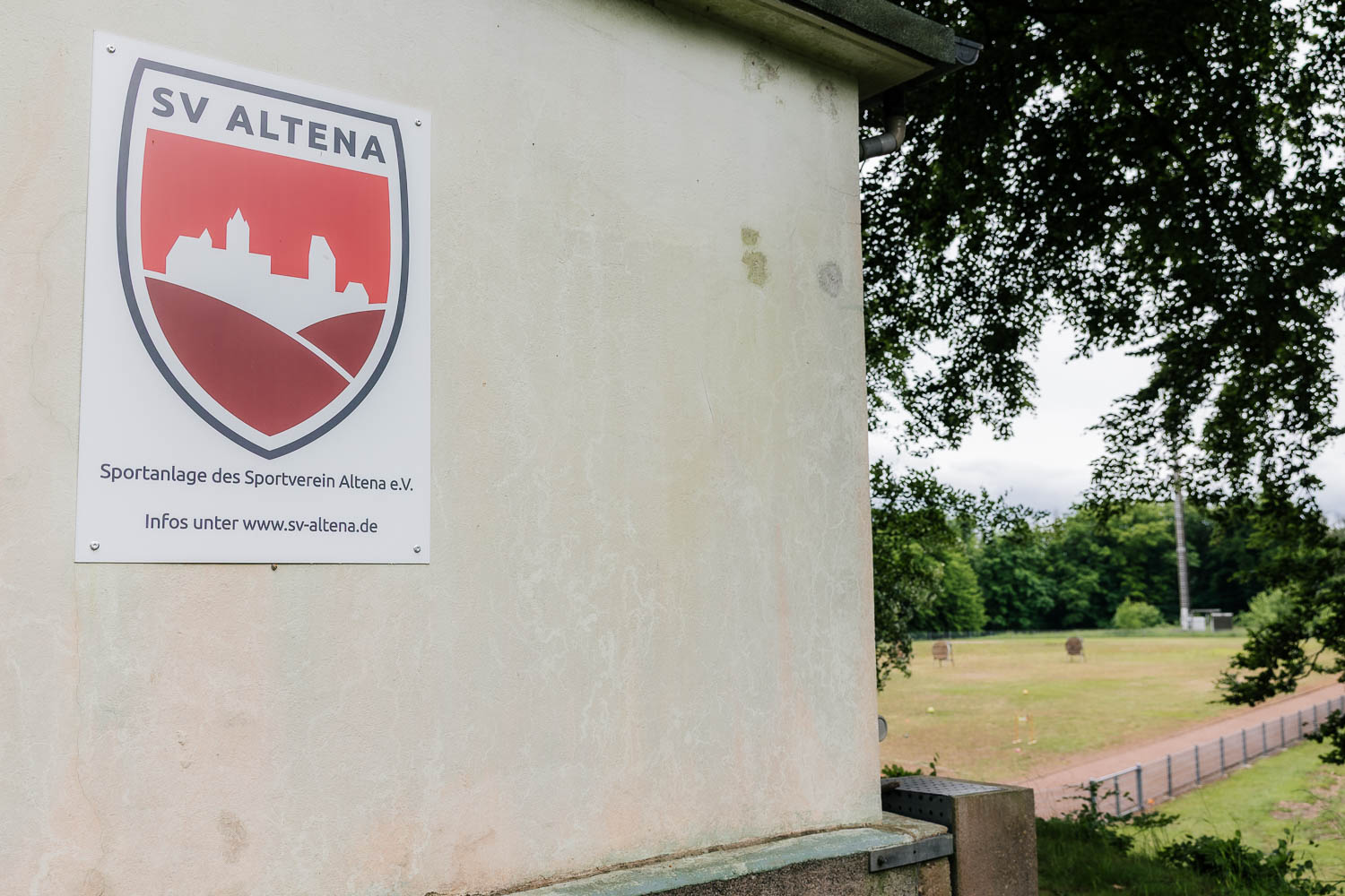 Das Bild zeigt links das Schild des SV Altena oben am Adolf-Hahn-Platz sowie rechts den Blick auf die Rasenfläche